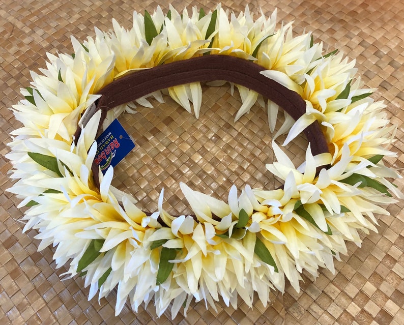 Puede incluir: Una vibrante corona de flores, con flores de color amarillo crema y blanco con detalles verdes. Las flores est&aacute;n dispuestas alrededor de una banda marr&oacute;n. Una etiqueta azul con texto est&aacute; adjunta. La corona se muestra sobre una superficie tejida.