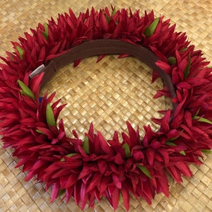 Puede incluir: Un lei floral rojo vibrante con acentos verdes y una banda marr&oacute;n. El lei est&aacute; hecho de numerosos p&eacute;talos rojos y hojas verdes, dispuestos en un patr&oacute;n circular. El lei es una guirnalda hawaiana tradicional.