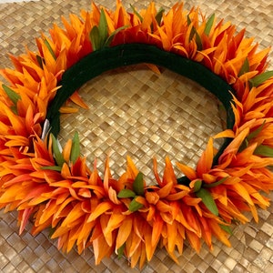Puede incluir: Una vibrante corona floral naranja con detalles verdes. La corona presenta capas de p&eacute;talos naranjas y hojas verdes, sobre una base verde oscuro. La corona se exhibe sobre una superficie tejida de color beige.