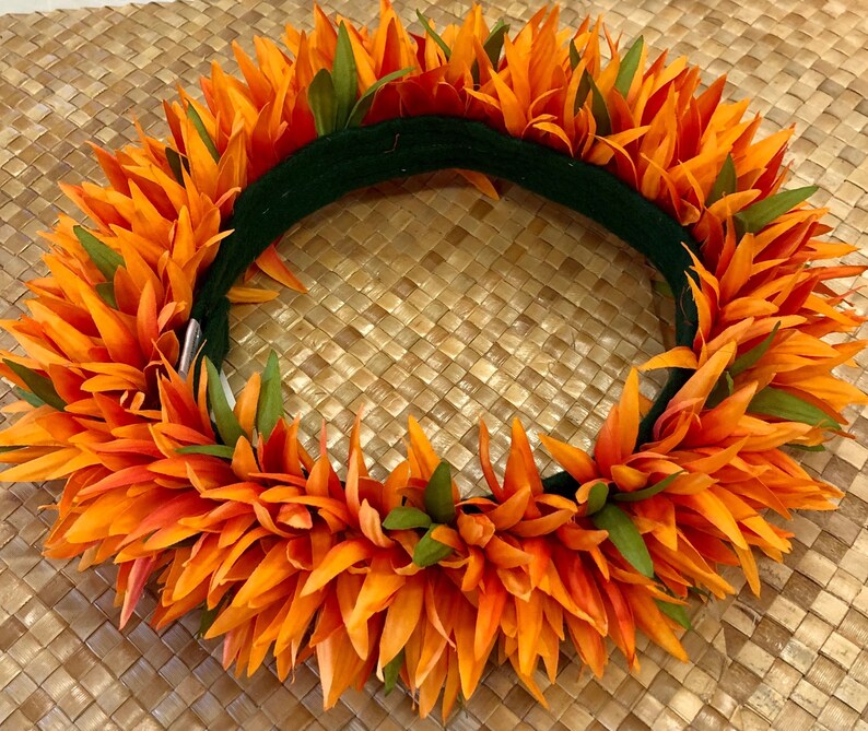 Puede incluir: Una vibrante corona floral naranja con detalles verdes. La corona presenta capas de p&eacute;talos naranjas y hojas verdes, sobre una base verde oscuro. La corona se exhibe sobre una superficie tejida de color beige.
