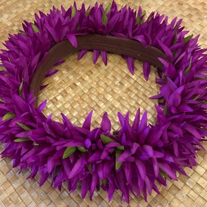 Puede incluir: Un vibrante lei floral p&uacute;rpura con detalles verdes. El lei est&aacute; hecho de p&eacute;talos puntiagudos en capas y tiene una banda marr&oacute;n. El lei se muestra sobre una superficie tejida de color marr&oacute;n claro.
