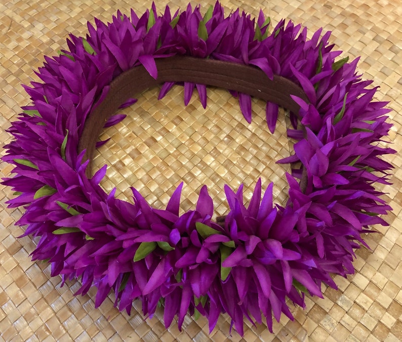 Puede incluir: Un vibrante lei floral p&uacute;rpura con detalles verdes. El lei est&aacute; hecho de p&eacute;talos puntiagudos en capas y tiene una banda marr&oacute;n. El lei se muestra sobre una superficie tejida de color marr&oacute;n claro.