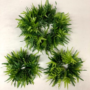 May include: Three artificial green fern wreaths of varying sizes. The wreaths are composed of multiple layers of green leaves, creating a full and textured appearance. The leaves have a pointed shape and a mix of light and dark green hues.