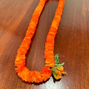 May include: An orange lei made of layered fabric, with a vibrant orange hue. The lei is adorned with a single orange hibiscus flower and green leaves. The lei is displayed on a dark wooden surface.