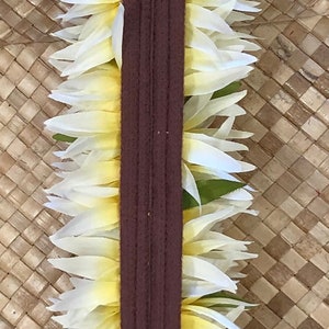 Puede incluir: Un lei, una guirnalda hawaiana tradicional, con p&eacute;talos de flores blancos y amarillos. El lei est&aacute; centrado en una banda de tela marr&oacute;n oscuro con cierre de velcro. El fondo es una estera tejida, marr&oacute;n claro. La etiqueta dice "Made in Hawaii."