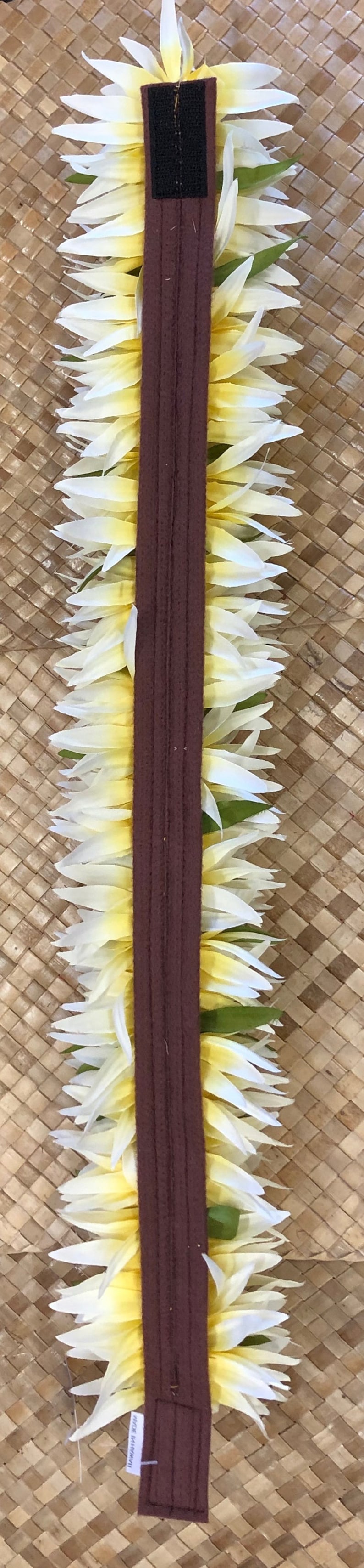 Puede incluir: Un lei, una guirnalda hawaiana tradicional, con p&eacute;talos de flores blancos y amarillos. El lei est&aacute; centrado en una banda de tela marr&oacute;n oscuro con cierre de velcro. El fondo es una estera tejida, marr&oacute;n claro. La etiqueta dice "Made in Hawaii."