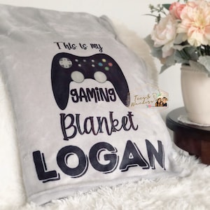 May include: A gray gaming blanket with a black controller graphic and the text "This is my GAMING BLANKET LOGAN". The blanket is made of soft material and is displayed on a white, fluffy surface.