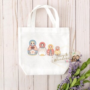 May include: White tote bag featuring a watercolor illustration of five Matryoshka dolls in various colors. The dolls are arranged in a row, with the largest on the left and the smallest on the right. The bag has two white handles.