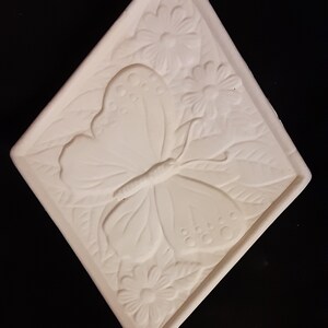 May include: A square, off-white ceramic tile with a raised butterfly and floral design. The butterfly is centered, with detailed wings and body. Flowers and leaves fill the background, creating a nature-inspired motif. The tile has a raised border.
