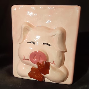 May include: A pink ceramic piggy bank with a 3D sculpted pig face. The pig has closed eyes and a pink snout. The pig is holding its right hoof to its mouth.