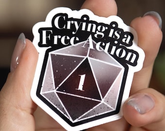 May include: A die-cut sticker featuring a twenty-sided die graphic in shades of dark purple and black with a white number one in the center. The text Crying is a Free Action is printed in a bold white font with a black outline above the die.