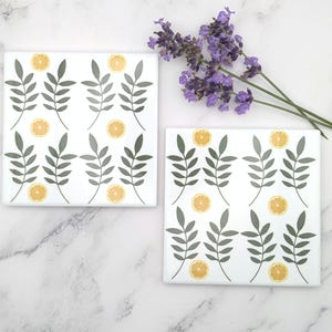 May include: Two square white coasters featuring a repeating pattern of dark green leafy branches and small yellow lemon slices. The coasters are arranged side by side with a sprig of purple lavender flowers placed near the top right corner.