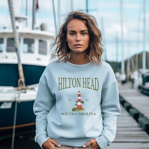 Può includere: Felpa blu chiaro con una grafica di un faro rosso e bianco con palme e il testo "HILTON HEAD" e "SOUTH CAROLINA".