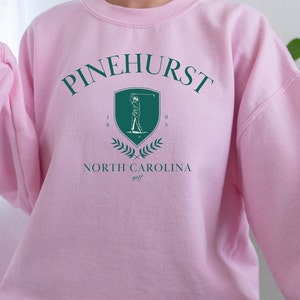 May include: Pink sweatshirt with a green graphic of a golfer and the text "Pinehurst North Carolina golf" with the numbers "1895" on the shield.