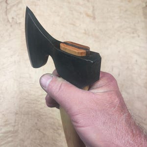 May include: A black axe head with a wooden handle. The axe head has a curved blade and a wooden accent piece on top. The handle is light-colored wood. The axe is held in a hand.