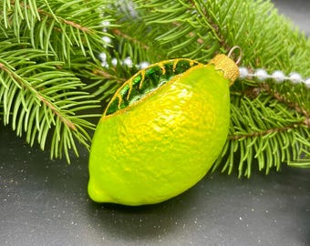 Op de afbeelding: Een kerstversiering in de vorm van een limoen met een getextureerd, heldergroen oppervlak en een goudkleurige dop. De versiering heeft een groen en goudkleurig bladerontwerp en ligt tegen een achtergrond van dennennaalden en witte kralen.