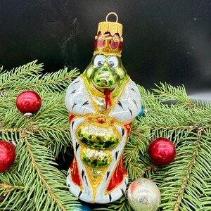 Op de afbeelding: Een glazen kerstornament in de vorm van een groen wezen met een gouden kroon en een wit gewaad met zwarte stippen en rode randen. Het figuur heeft een rode tong, grote witte ogen en een gouden ophanglus aan de bovenkant van de kroon.