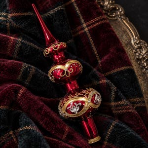 May include: A shiny red glass Christmas tree topper featuring a pointed spire, two rounded bulbous sections, and intricate gold filigree detailing. The surface is embellished with small clear crystals arranged in floral patterns across the base.