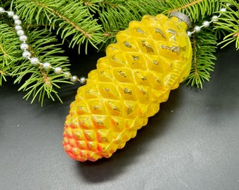 May include: A vintage Christmas ornament shaped like a pine cone. The ornament is yellow with gold accents and a red-orange tip. It rests on a dark surface with evergreen branches and a silver beaded garland.