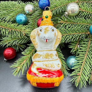 Op de afbeelding: Een glazen kerstornament in de vorm van een cobra met een gele kroon. De slang is wit met gouden glitteraccenten, een rode tong en rust op een rood-gele basis. Het ornament is voorzien van een gouden ophanglus aan de bovenkant van de kroon.