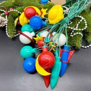 May include: A selection of retro Christmas lights in assorted shapes and colours, including red, blue, yellow, and green. The lights are strung on green wire and are displayed with evergreen branches and decorative beads.