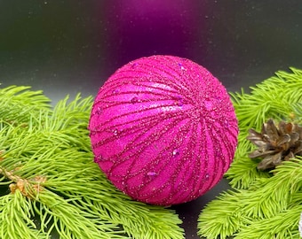 May include: A magenta Christmas ornament with a glittery, textured surface and a radial design. The ornament is surrounded by green pine needles and a pine cone against a black background, creating a festive holiday scene.