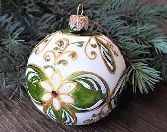 May include: White Christmas ornament with green and gold floral designs. The ornament has a red jewel in the center of the flower.