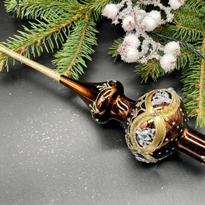 Könnte beinhalten: Ein braun-goldener Weihnachtsbaumaufsatz mit aufwendigen Details und funkelnden Akzenten. Der dekorative Glasschmuck hat eine lange, glitzernde goldene Spitze und ist vor einem Hintergrund aus Tannenzweigen platziert.