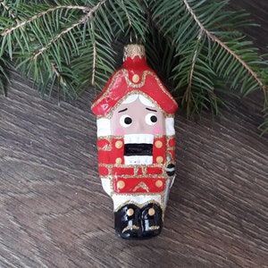 May include: A red and white glass Christmas ornament shaped like a nutcracker soldier. The ornament has a gold glitter finish and black painted details.