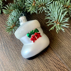 May include: A white Christmas boot ornament with a black sole, adorned with green and red bells and bows. The ornament has a silver hanger and is set against a wooden background with pine branches.
