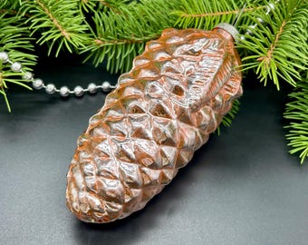 May include: A vintage Christmas ornament shaped like a pine cone. The ornament is a metallic copper color with a textured surface. The background includes green pine branches and a string of silver beads.