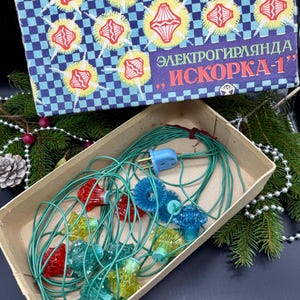 Könnte beinhalten: Vintage Weihnachtsbeleuchtung in einer Pappschachtel. Der Deckel der Schachtel hat ein blau-weißes Karomuster mit Sternenmustern und Text. Die Lichter sind in verschiedenen Farben gehalten, darunter Rot, Blau und Gelb, mit grünen Kabeln.