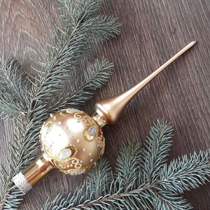 Op de afbeelding: Een gouden kerstboomtopper met een ronde, versierde bal en een lange, puntige toren. De topper is omringd door groene dennentakken.