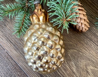 May include: A gold glass Christmas ornament shaped like a pine cone. The ornament has a textured surface and is hanging from a green pine branch.