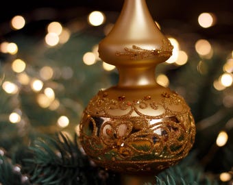 May include: A gold Christmas tree topper with a decorative, ornate design. The topper is set against a backdrop of blurred lights and evergreen branches, with silver beads. The topper has a bulbous base and a pointed top.