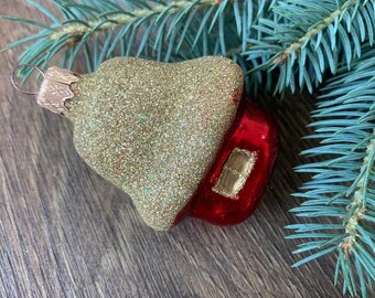 May include: A vintage Christmas ornament shaped like a house, with a gold glitter top and a red base. The ornament has a small window and a metal cap for hanging. The background includes blue-green pine branches.