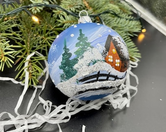 May include: A hand-painted Christmas ornament with a winter scene. The blue glass ball features a snow-covered landscape with a cabin, trees, and a fence. The ornament is accented with glitter and sits among evergreen branches.