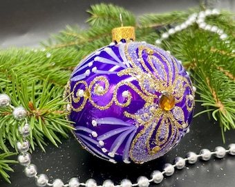 May include: A purple Christmas ornament with gold and white decorative accents. The ornament has a gold cap and a large, amber-colored jewel. The ornament is surrounded by pine needles and a string of silver beads.