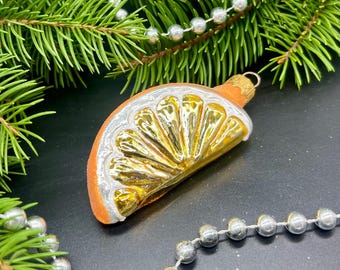 May include: A vintage Christmas ornament shaped like a slice of orange. The ornament has a gold interior, silver rind, and orange exterior. The ornament is hanging from a gold metal loop. The background includes pine needles and silver bead garland.