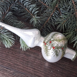 May include: A white glass Christmas tree topper with a green and red holly design. The topper is shaped like a teardrop with a pointed top.