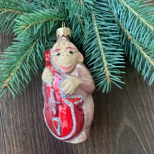 Puede incluir: Un adorno de Navidad de vidrio con forma de mono tocando un violonchelo rojo. El mono es marrón con la cara blanca y los ojos negros. El adorno tiene una parte superior dorada para colgar.