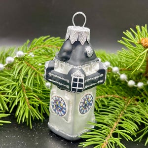 Puede incluir: Adorno navideño con forma de torre del reloj. El adorno es blanco con detalles negros y adornos de purpurina plateada. Las esferas del reloj tienen detalles azules, amarillos y rojos. Cuelga de un lazo plateado y está rodeado de agujas de pino verdes.