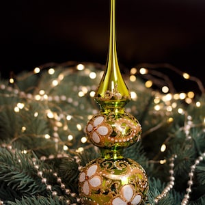Op de afbeelding: Een groene en gouden kerstboomtop met een lange, puntige top. De topper heeft twee decoratieve bollen met witte bloemmotieven en gouden accenten. De achtergrond bevat een kerstboomtak, een parelsnoer en een lichtslinger.