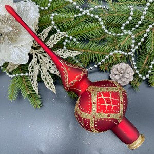 Puede incluir: Un adorno rojo para la parte superior del árbol de Navidad con detalles dorados. El adorno tiene un tallo largo y un cuerpo bulboso con un diseño cuadrado dorado. El fondo incluye ramas de pino, cuentas plateadas y una flor decorativa.