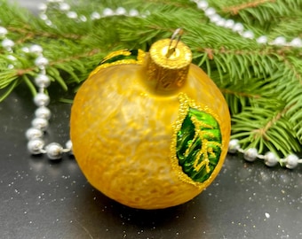 Puede incluir: Una bola de Navidad redonda, de color amarillo dorado, con un diseño de hoja verde y detalles dorados. El adorno cuelga frente a una rama de pino verde y una guirnalda de cuentas plateadas.