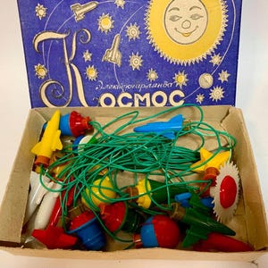 Op de afbeelding: Een vintage kerstverlichtingsset in een kartonnen doos. Het deksel van de doos is blauw met illustraties van een lachende zon en raketten. De lampjes hebben de vorm van raketten, sterren en andere ruimtevaartthema's, verbonden door een groene draad.