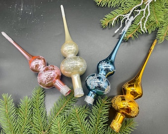 May include: Four colorful glass Christmas tree toppers. The toppers are in shades of red, gold, blue, and champagne. Each has a long, pointed tip and a decorative, round base with snowflake designs. Glitter accents the bases.
