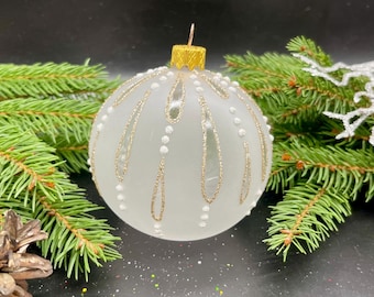 May include: A frosted white Christmas ornament with gold glitter accents and white beaded details. The ornament has a gold cap and is surrounded by green pine branches and a pine cone. The background is black.