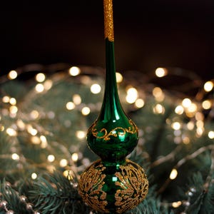 Könnte beinhalten: Ein grüner Weihnachtsbaumaufsatz mit goldenen Glitzerakzenten. Der Aufsatz hat eine bauchige Basis und eine schlanke, längliche Spitze. Goldene Filigranarbeiten verzieren die Basis und die Spitze. Der Hintergrund zeigt einen Weihnachtsbaum und Lichterketten.