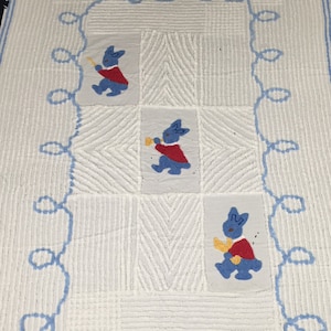 May include: White crocheted baby blanket with a blue border and three blue and red bunny appliques holding a horn.